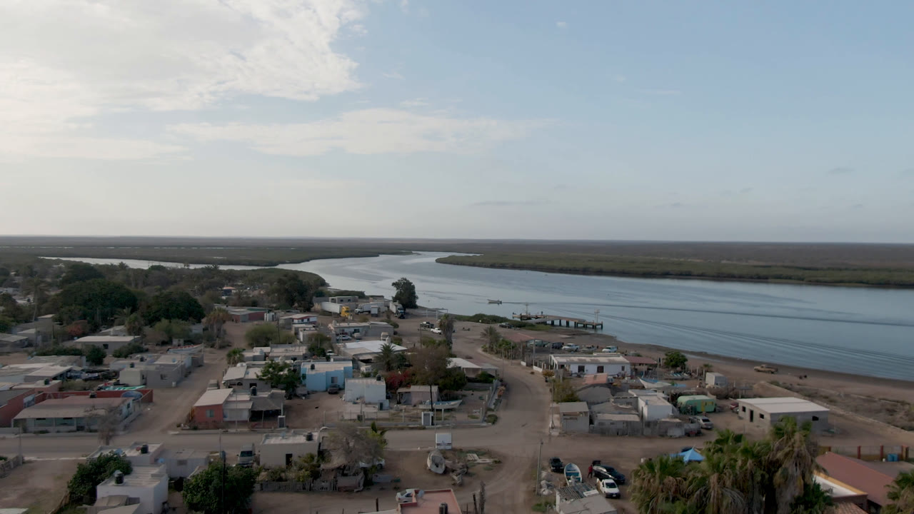 Aerial view in Adolfo Lopez Mateos, Mexico, with the settlement filling the lower frame, dock structures near the shore, open water across the middle, mangroves on the far bank, and no visible people.