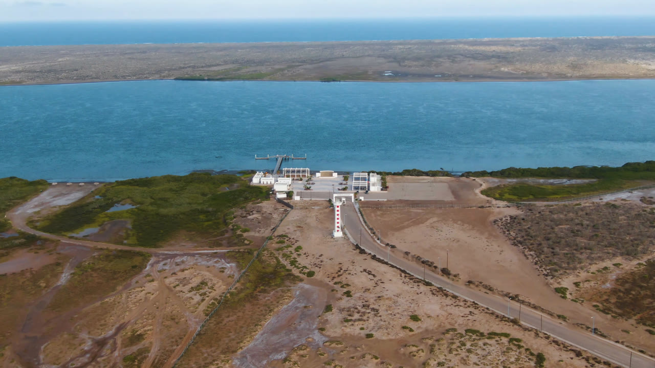 High aerial frame over Aldofo Lopez Mateos, Baja California Sur, showing a white coastal building, a long walkway through sandy scrubland, blue water beyond, and no visible people.