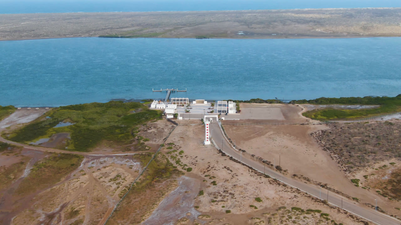 Aerial frame in Aldofo Lopez Mateos with the walkway leading toward the white shoreline building, open sand and scrub on both sides, blue water behind it, and no visible people.
