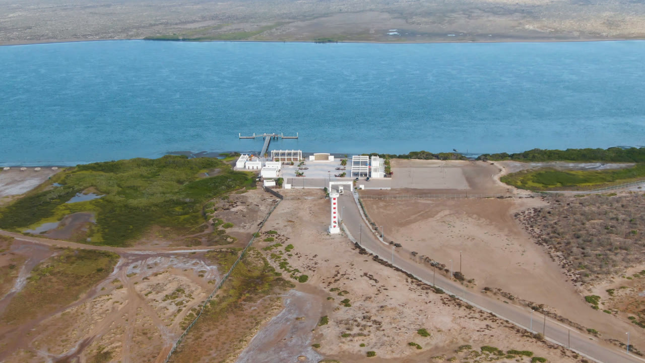 Wide drone frame of a coastal building beside turquoise water and dry terrain.