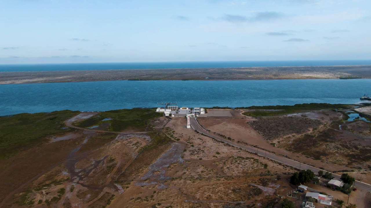Wide aerial frame over Aldofo Lopez Mateos, Baja California Sur, showing a long road leading toward a white waterfront building, blue water, low shoreline, scrub terrain, and no visible people.