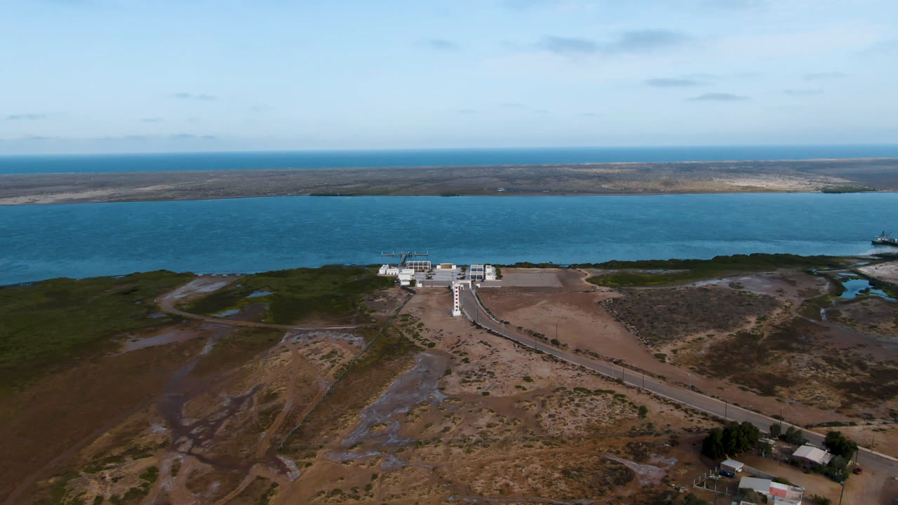 Aerial view in Aldofo Lopez Mateos with the road running from the lower frame toward the waterfront building, dark green vegetation near the shore, blue lagoon water, and no visible people.
