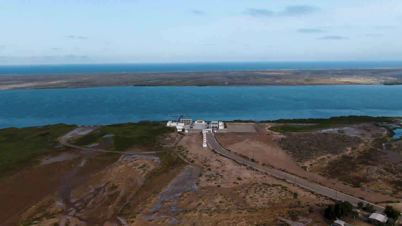 Wide coastal aerial with building, road, water, and distant low land.