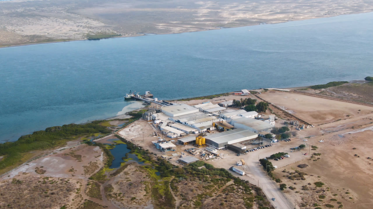 Opening aerial frame over Comondu, Mexico, showing a large factory complex near blue water, arid scrubland, sandy access roads, and no visible people.