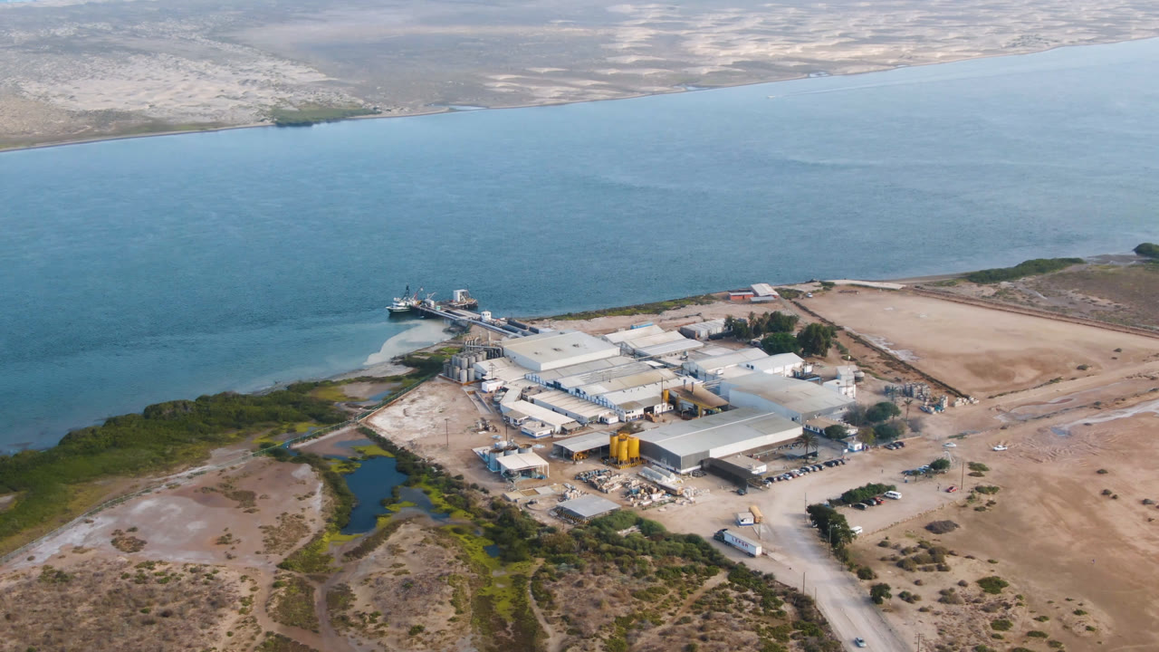 Aerial frame in Comondu, Mexico, with the factory centered below the horizon, shoreline water behind it, a small wetland channel at left, and no visible people.