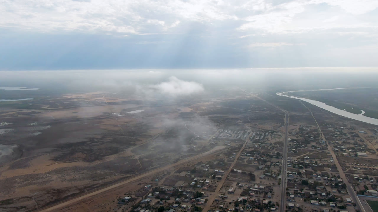 High aerial view over Aldofo Lopez Mateos, Baja California Sur, with town blocks below, a river in the distance, drifting haze, clouded daylight, and no visible people.