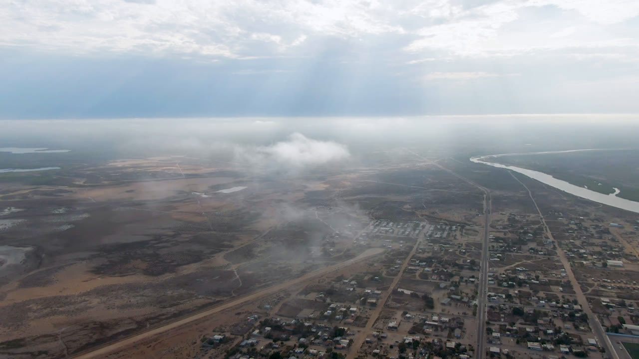 Second aerial frame over Aldofo Lopez Mateos shows the central road axis, clustered low buildings, a pale river bend at right, hazy air, and no visible people.
