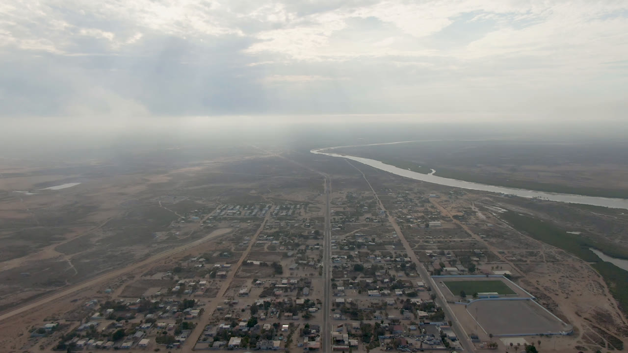 High aerial frame from Aldofo Lopez Mateos showing clustered buildings at the bottom, a straight road leading toward the horizon, a winding river in the midground, and no visible people.