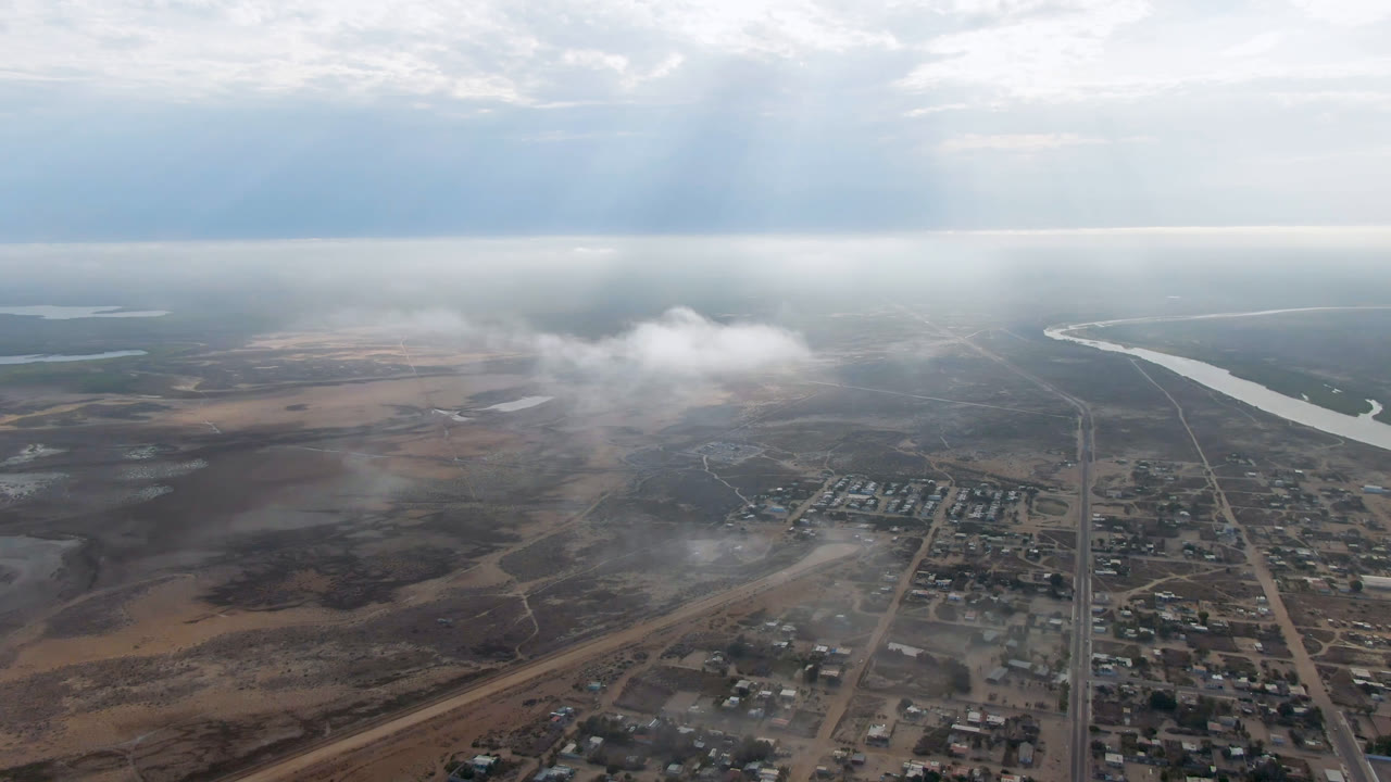 High aerial view over Aldofo Lopez Mateos, Baja California Sur with town blocks below, a winding river in the distance, haze across the sky, and no visible people.