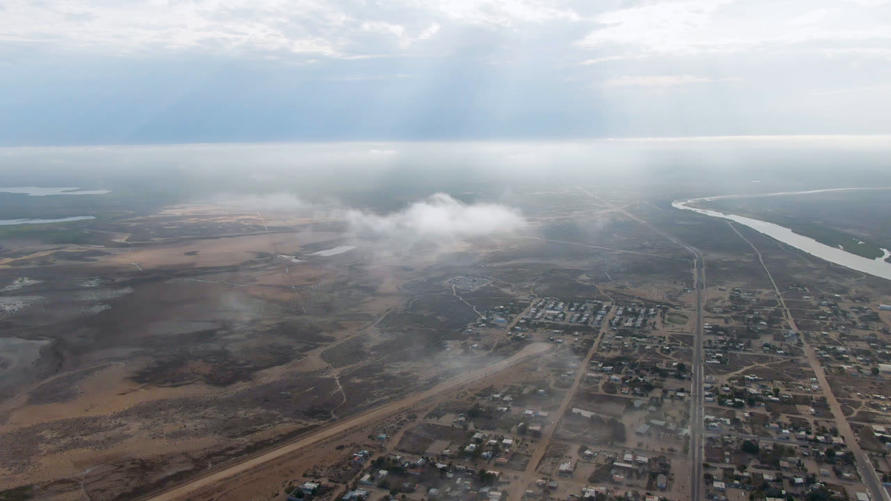 Aerial frame over Aldofo Lopez Mateos showing a straight road corridor through low buildings, brown open land, a distant river bend, haze, and no visible people.