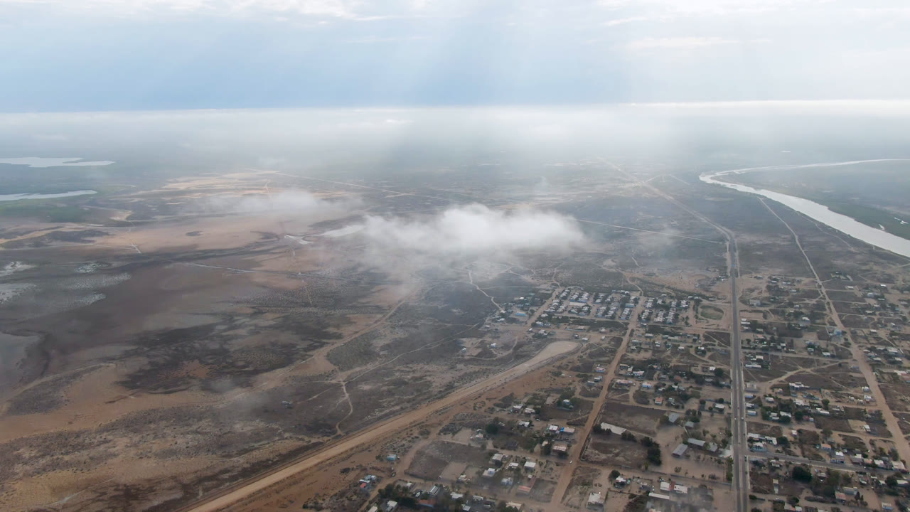Aerial frame over Aldofo Lopez Mateos showing the town blocks lower right, dry open terrain to the left, a bright winding river near the horizon, haze, and no visible people.
