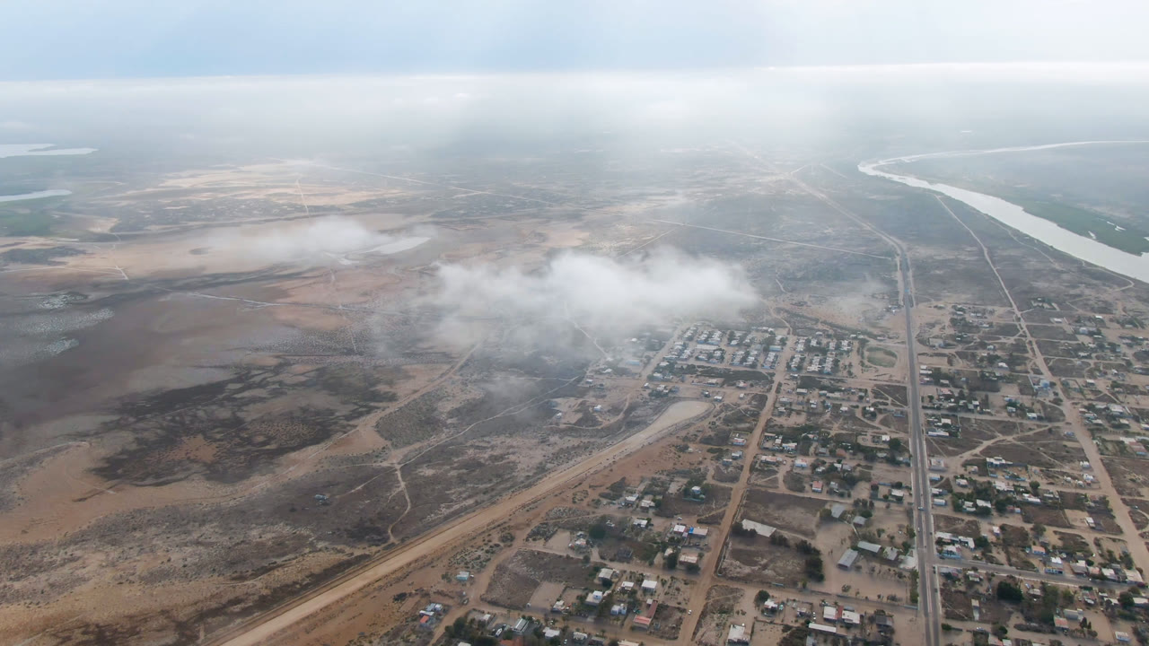 Aerial view of a dry town grid with roads, buildings, haze, and a river in the distance.