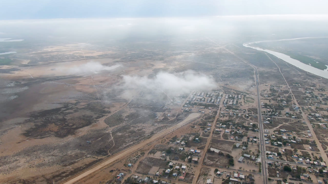 Aerial frame over Aldofo Lopez Mateos with dust or haze crossing the center, compact building blocks below, dry open land to the left, river water near the horizon, and no visible people.
