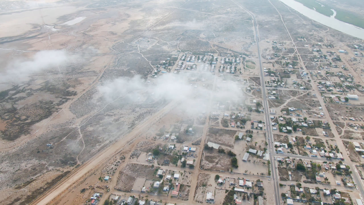 Aerial view over Aldofo Lopez Mateos, Baja California Sur with a vertical road dividing buildings and open terrain while white dust or cloud softens the middle of the frame, no visible people.