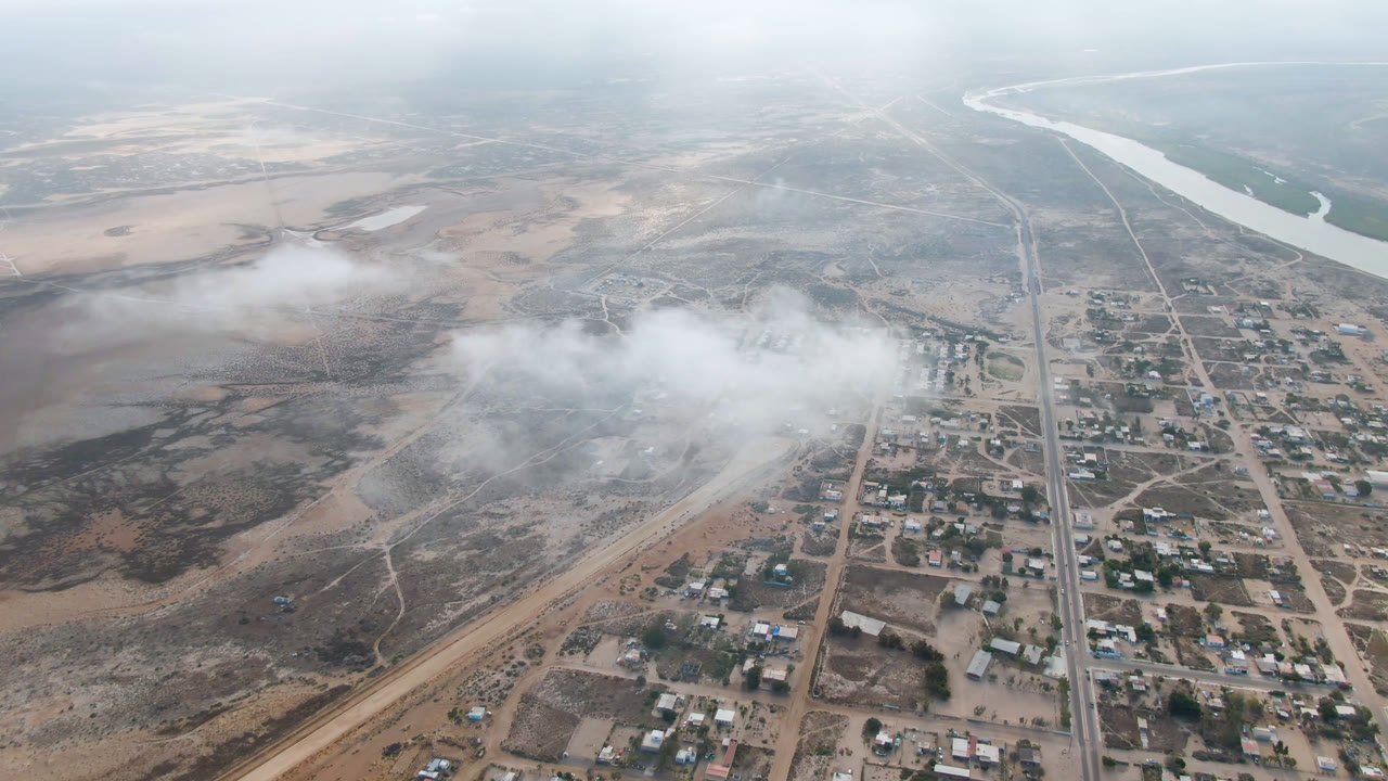 Aerial frame from Aldofo Lopez Mateos with a central road grid, small buildings, scrub-covered ground, drifting haze across the lower middle, distant river water, and no visible people.