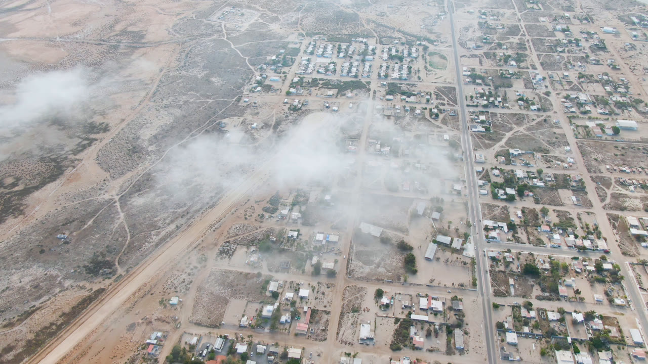 High aerial frame over Aldofo Lopez Mateos, Baja California Sur, showing pale dirt roads, low buildings, scattered trees, and white haze drifting across the left side, with no visible people.