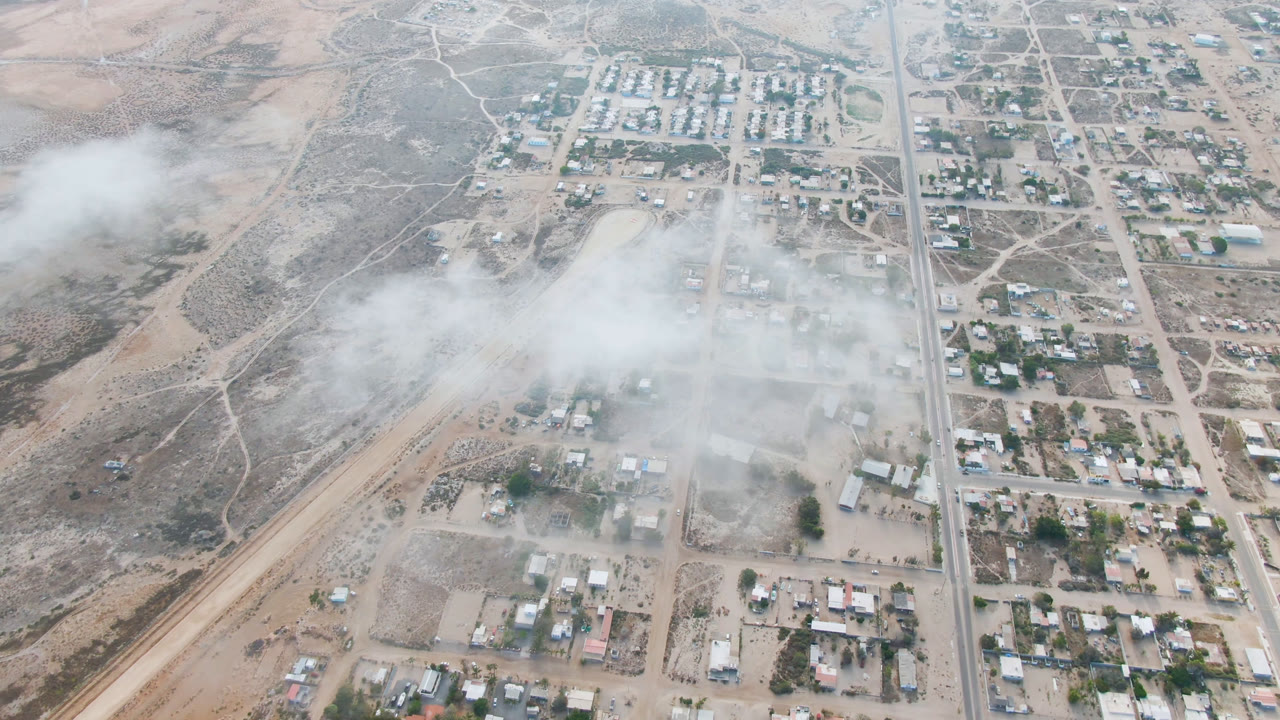 Aerial view of Aldofo Lopez Mateos with a straight road near the right side, compact town blocks, open desert ground, and haze covering the central buildings, with no visible people.