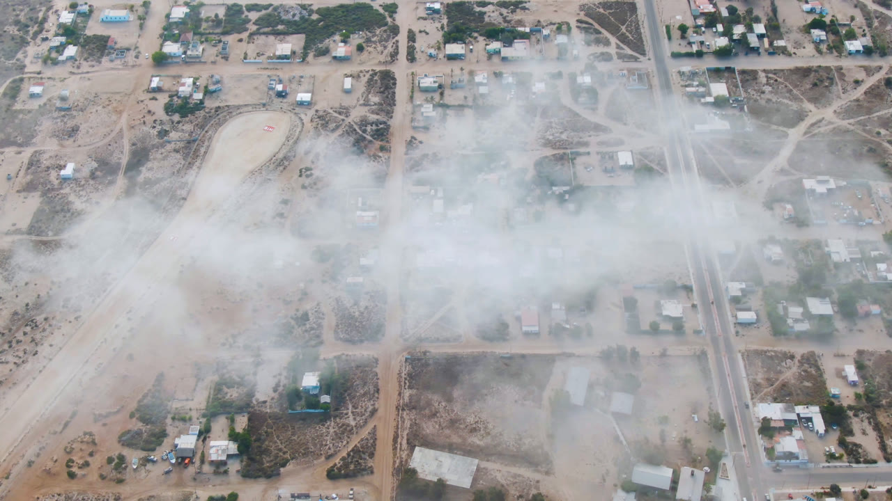 Overhead aerial frame in Aldofo Lopez Mateos, Baja California Sur, showing a wide pale smoke band over sandy roads, scattered buildings, vehicles, trees, and no visible people.