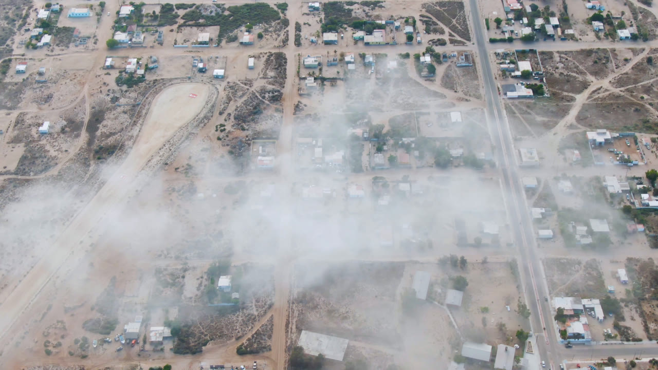 Overhead aerial frame of Aldofo Lopez Mateos, Baja California Sur, with tan roads, scattered buildings, sparse trees, parked vehicles, and a pale smoke or dust band across the center, no visible people.
