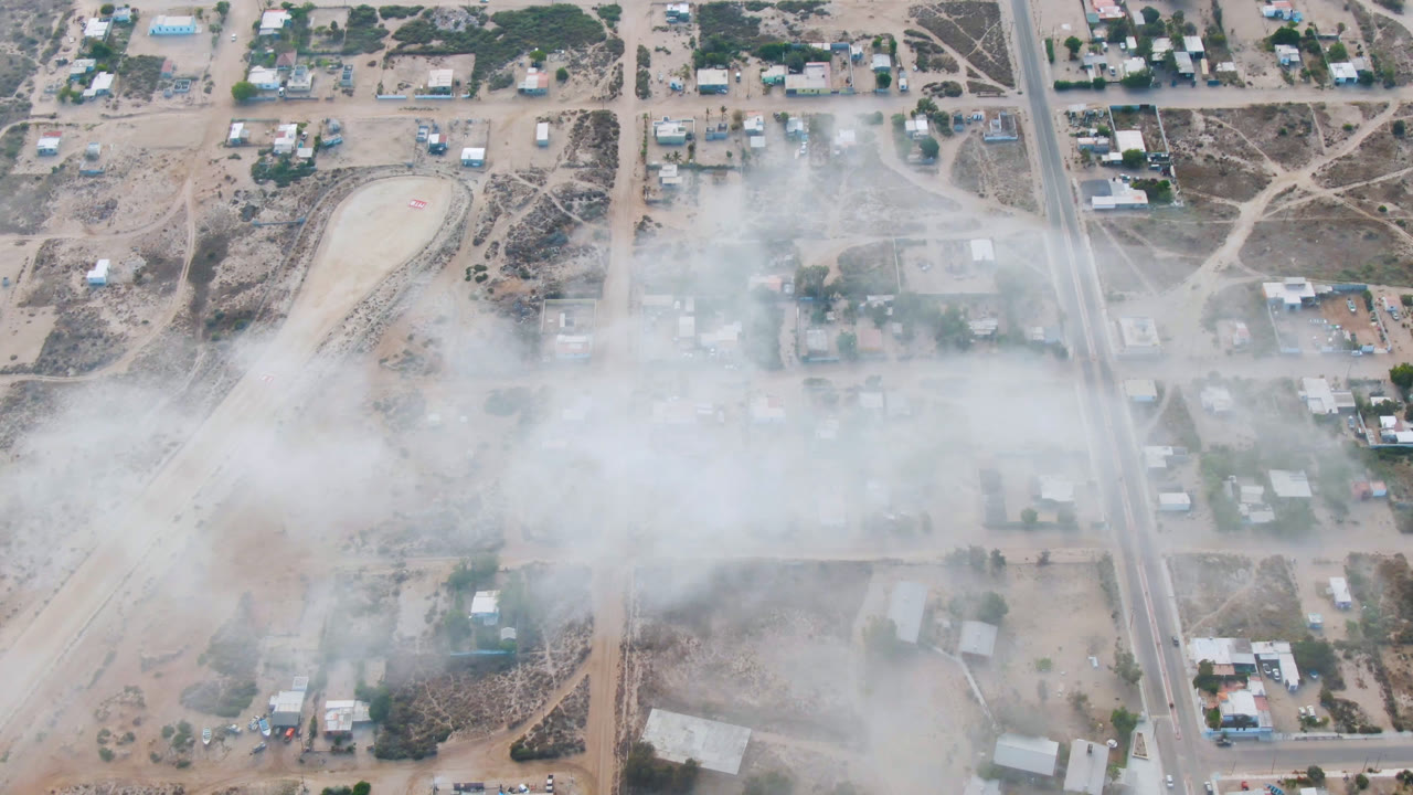Aerial view over Aldofo Lopez Mateos showing a straight road, low buildings, dry lots, and smoke or dust thickening across the mid-frame, with no visible people.