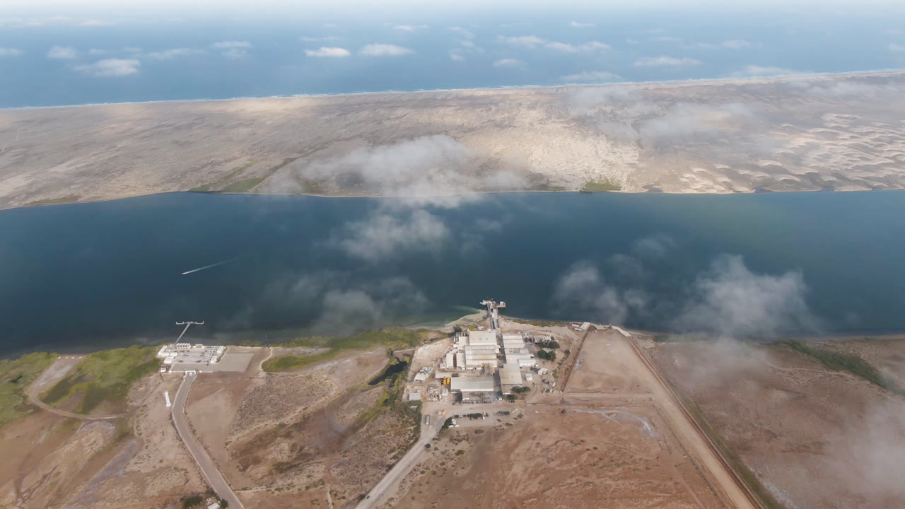 High aerial frame over Aldofo Lopez Mateos, Baja California Sur, with dark water beyond a desert shoreline, low clouds, roads, an industrial complex near the lower center, and no visible people.