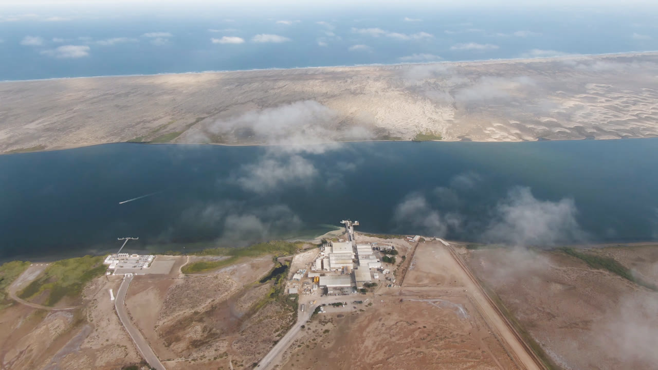 Aerial view in Aldofo Lopez Mateos, Baja California Sur, showing a shoreline industrial site below, dark blue water through the middle, pale desert land beyond, cloud patches, and no visible people.