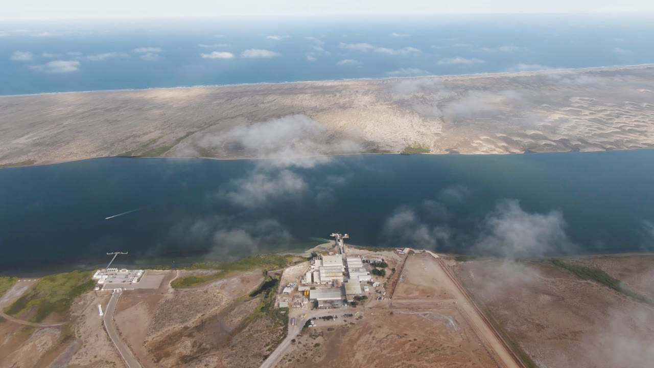 Wide drone view of water, desert flats, scattered clouds, and shoreline structures near cleared ground.