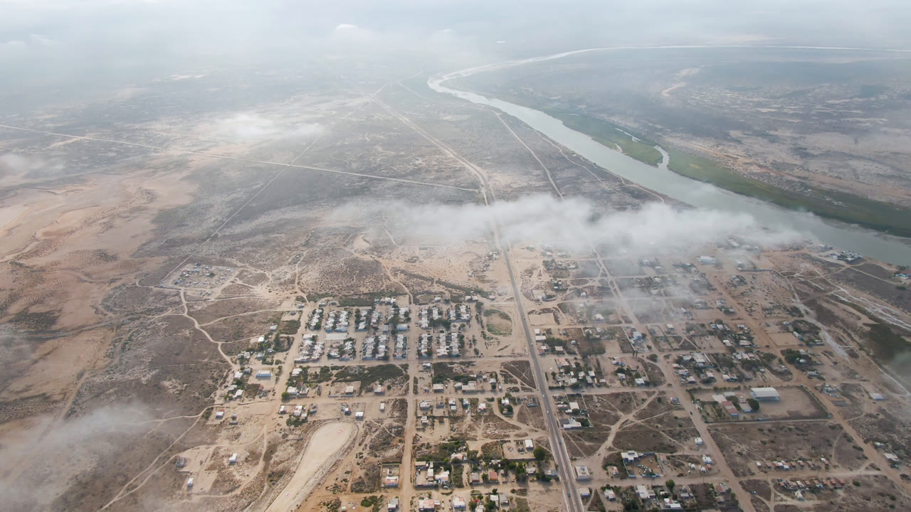 High aerial view over Aldofo Lopez Mateos, Baja California Sur, with a small town grid below, a river near the horizon, dry terrain around it, and no visible people.