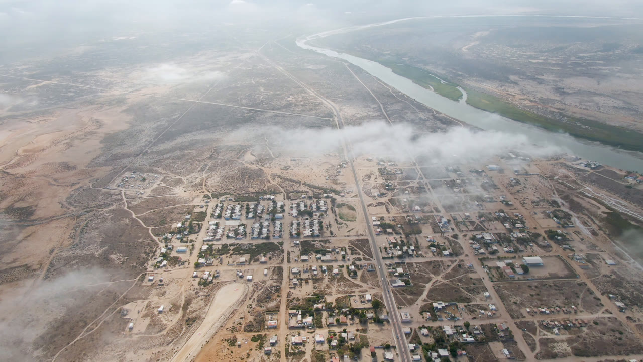 Aerial frame of Aldofo Lopez Mateos showing straight roads, clustered buildings, pale desert ground, drifting cloud cover, and the river running across the upper frame with no visible people.