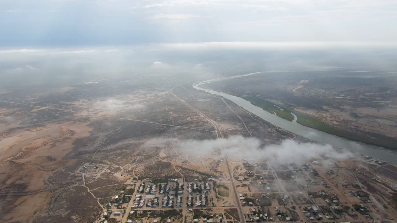 Opening aerial frame over Aldofo Lopez Mateos, Baja California Sur, with a curving river, gridded town blocks, dry terrain, low clouds, and no visible people.