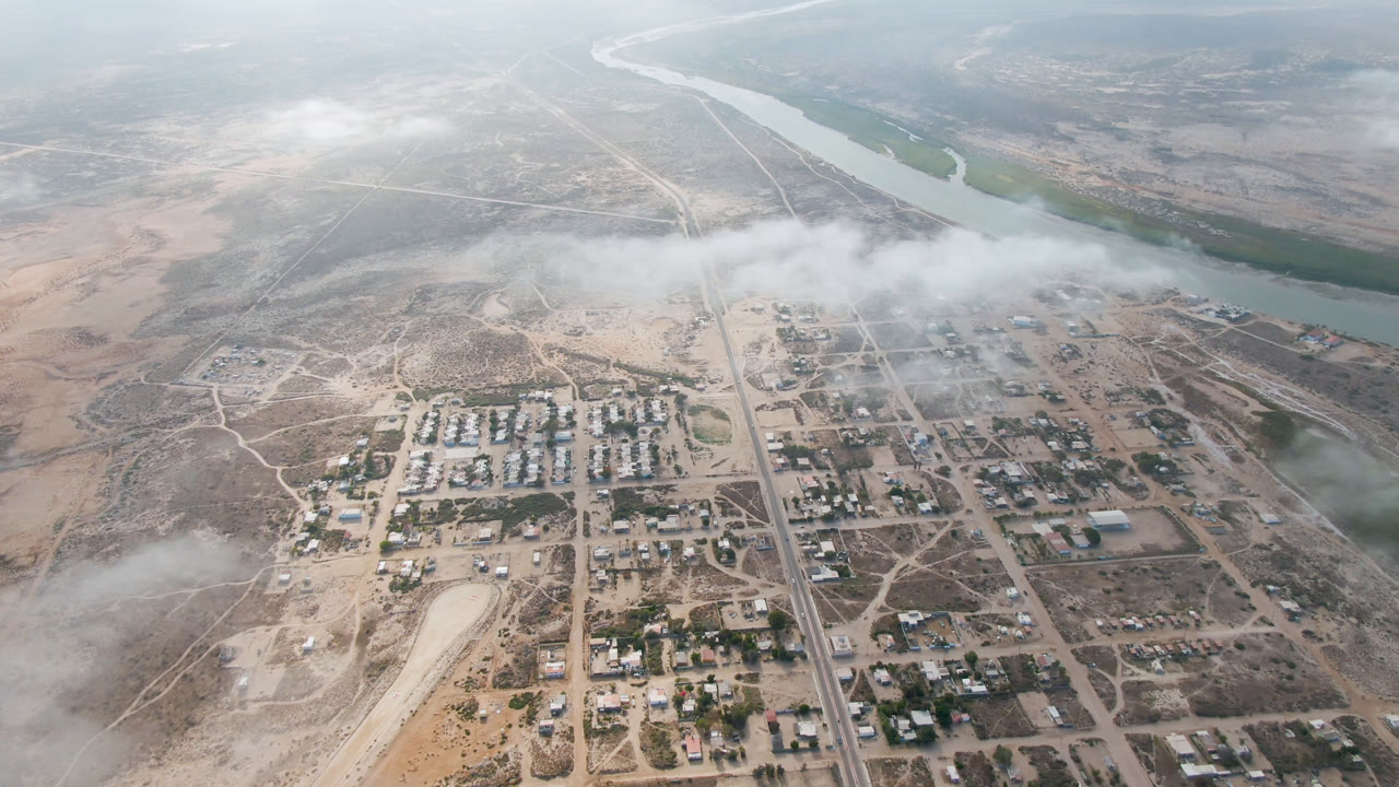 High aerial frame over Aldofo Lopez Mateos, Baja California Sur, with a town grid, pale desert ground, a curving river in the background, low cloud cover, and no visible people.