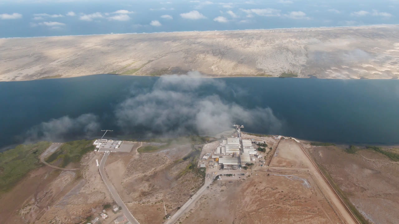 Aerial view of blue water, dry hills, roads, industrial buildings, and smoke.