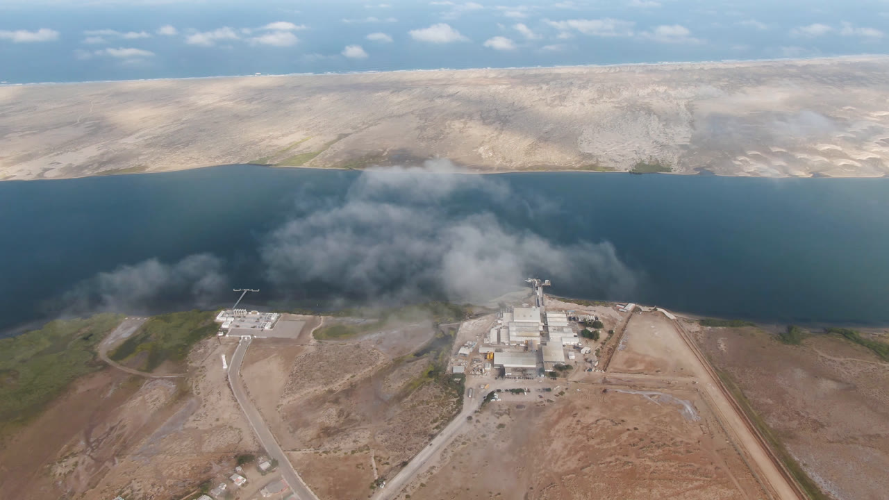 Wide aerial view in Aldofo Lopez Mateos, Baja California Sur, where smoke spreads above the near shore of a blue water body and industrial buildings sit below, no visible people.