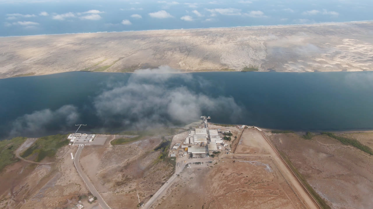 Aerial frame over Aldofo Lopez Mateos, Baja California Sur, showing an industrial facility near a broad dark water body, tan shoreline hills, scattered clouds, and no visible people.