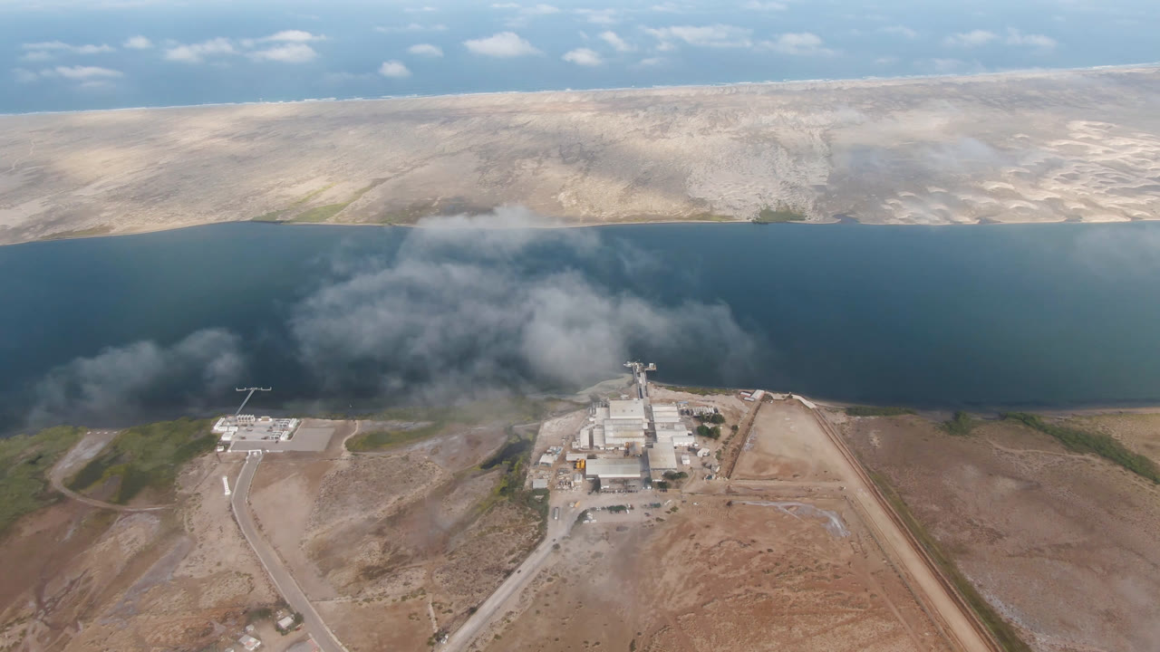 Drone view in Aldofo Lopez Mateos, Baja California Sur, with a white industrial complex near the shore, dirt roads in the foreground, dark water across the middle, distant arid hills, and no visible people.