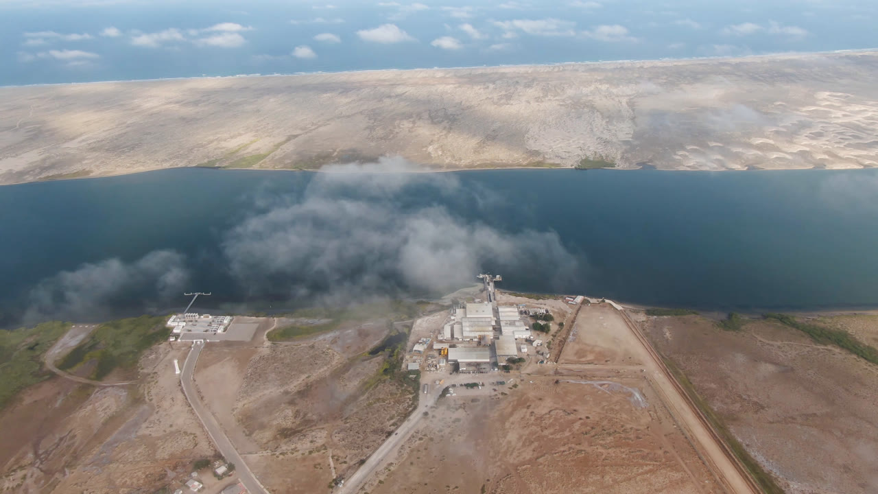 Elevated view of a shoreline factory with a cloud or smoke plume over water.