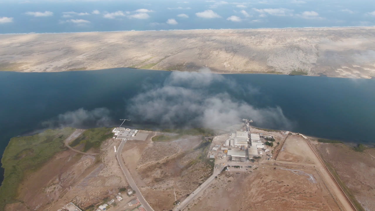 Aerial frame over Aldofo Lopez Mateos, Baja California Sur, showing blue water, pale hills, a shoreline building complex, roads, and no visible people at 0:00.