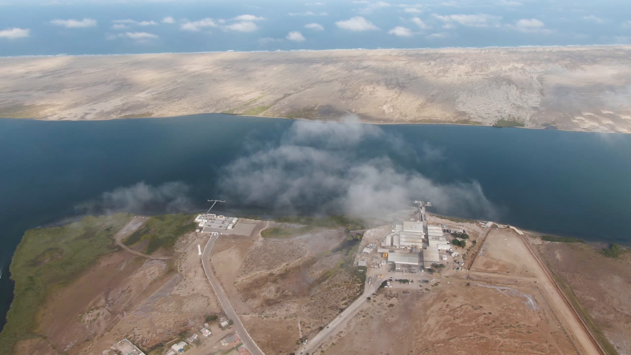 Aerial frame over Aldofo Lopez Mateos, Baja California Sur, with low cloud or smoke drifting over blue water behind a small shoreline industrial site and no visible people.