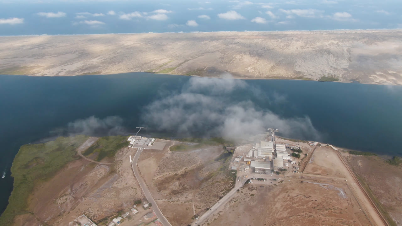 Aerial shoreline scene with blue water, pale hills, roads, and buildings below.