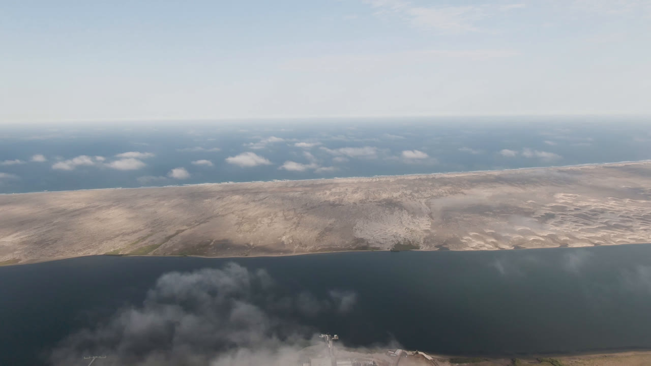 Opening aerial frame over Aldofo Lopez Mateos, Baja California Sur, with dark water below, pale desert shoreline across the middle, blue sky above, and no visible people.