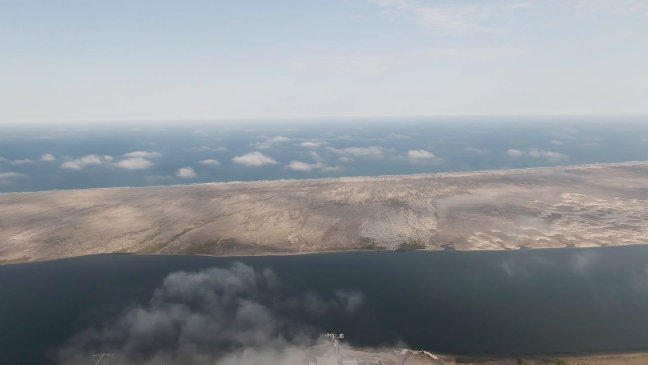 Aerial frame in Aldofo Lopez Mateos, Baja California Sur, showing a broad band of water, sandy terrain, small clouds near the horizon, and no visible people.
