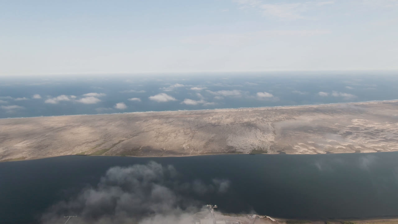 Aerial coastline view with dark water, pale land, distant horizon, and gray plume below.