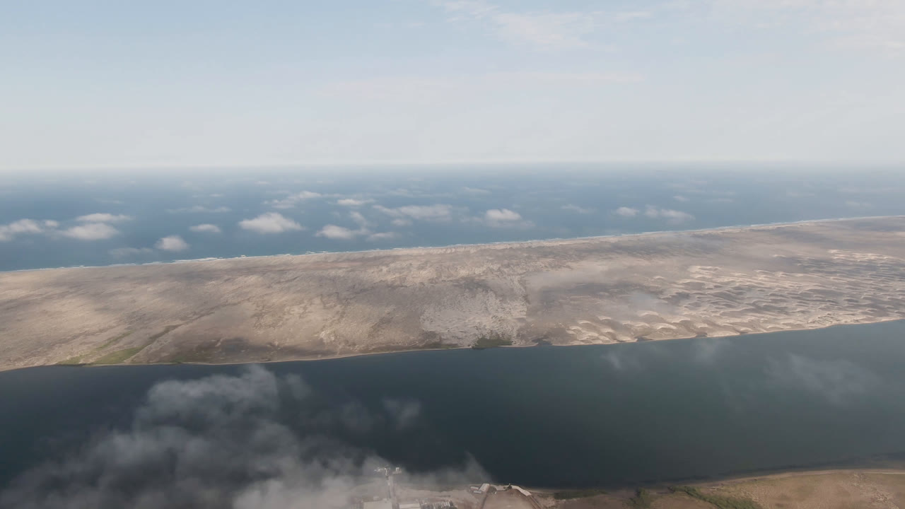 Opening aerial frame over Aldofo Lopez Mateos, Baja California Sur, with dark water in the foreground, pale dry land beyond it, low haze near the horizon, and no visible people.