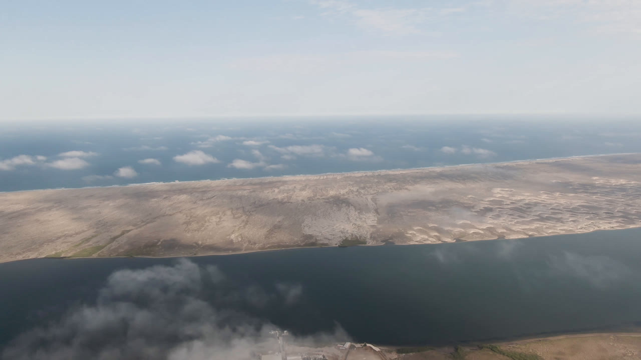Second aerial frame over Aldofo Lopez Mateos, Baja California Sur, with the water band crossing the lower frame, dry terrain across the middle, scattered clouds above, and no visible people.