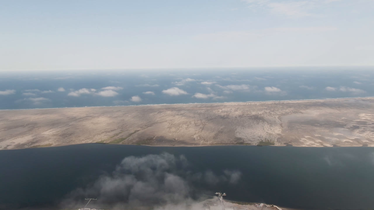 Aerial frame over Aldofo Lopez Mateos, Baja California Sur, Mexico, showing dark water below, pale sandy land across the middle distance, low horizon clouds, gray smoke near the lower shoreline, and no visible people.