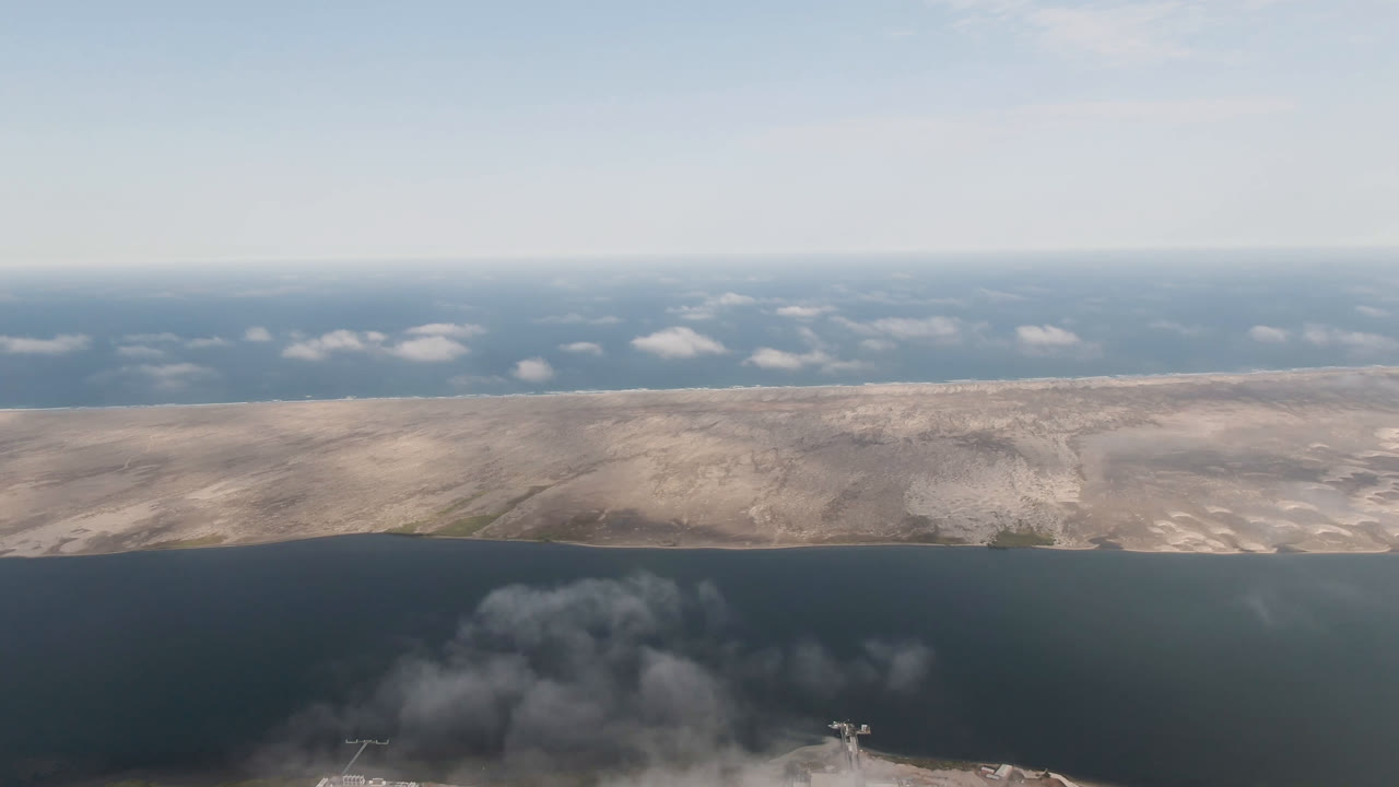 Aerial view in Aldofo Lopez Mateos, Baja California Sur, Mexico, with the smoke plume slightly more centered below the water, pale land stretching across the frame, a flat horizon, and no visible people.