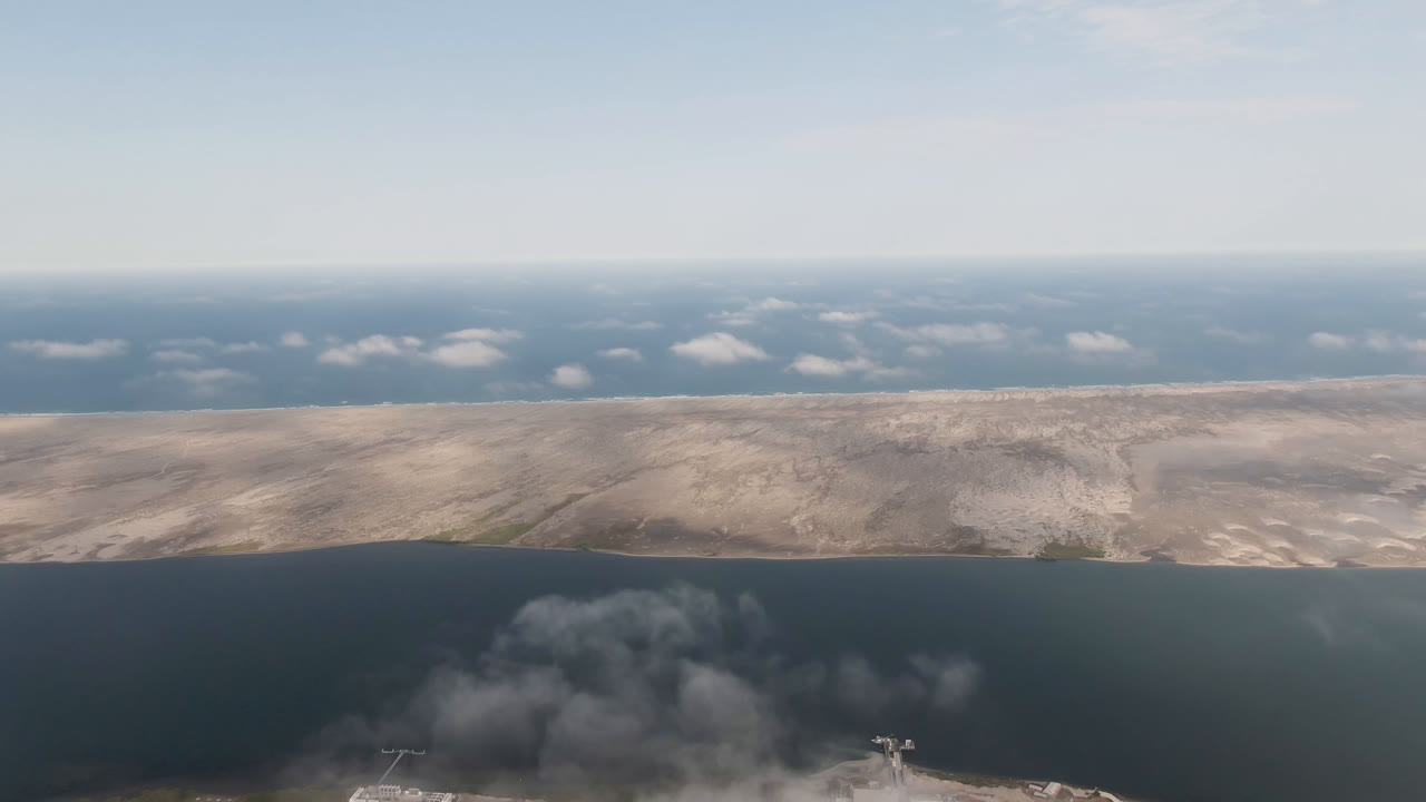 Aerial coastal view with dark water, sandy island-like land, distant clouds, and gray smoke below.