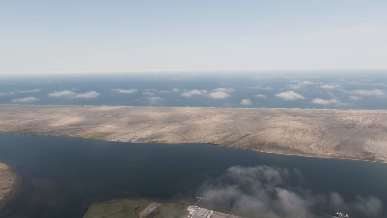Aerial frame over Aldofo Lopez Mateos, Baja California Sur, showing dark water, pale dry land, a low foreground island edge, scattered clouds, and no visible people.