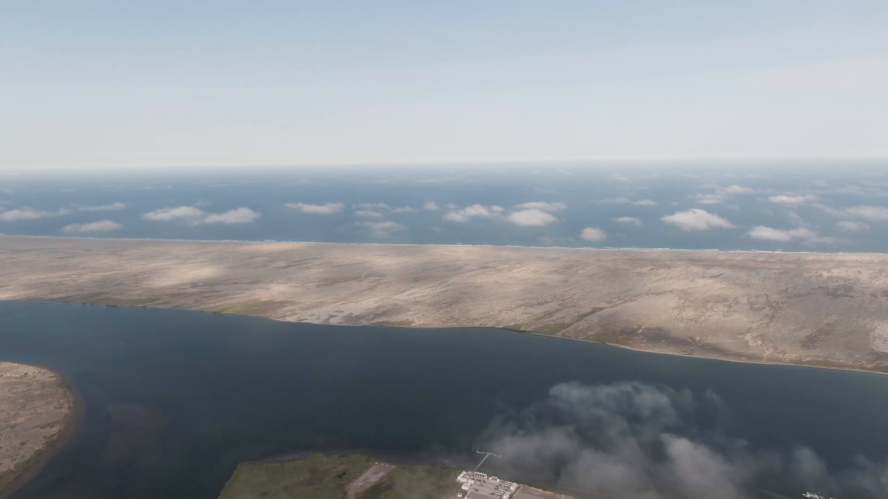 Second frame over Aldofo Lopez Mateos, Baja California Sur, with the water channel curving around scrubland, distant beige landforms, a clear horizon, and no visible people.