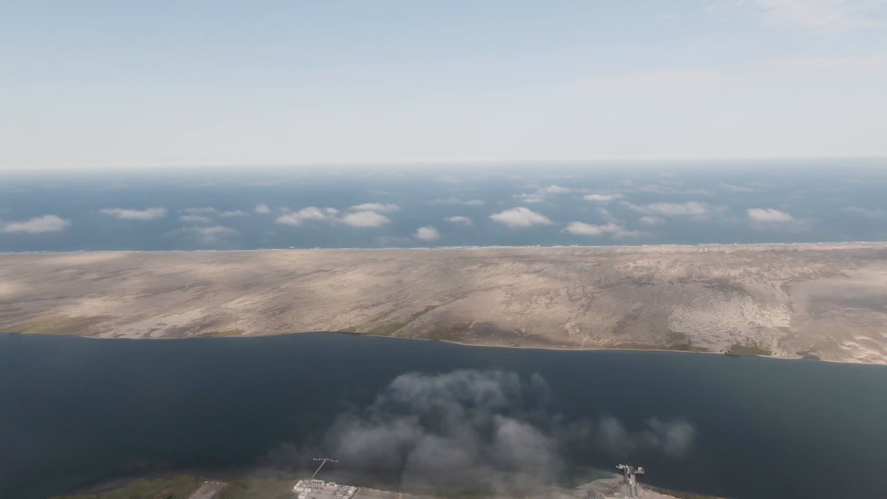 Aerial frame over Aldofo Lopez Mateos, Baja California Sur, Mexico, with dark water in the foreground, a pale dry shoreline across the middle, low clouds near the horizon, and no visible people.